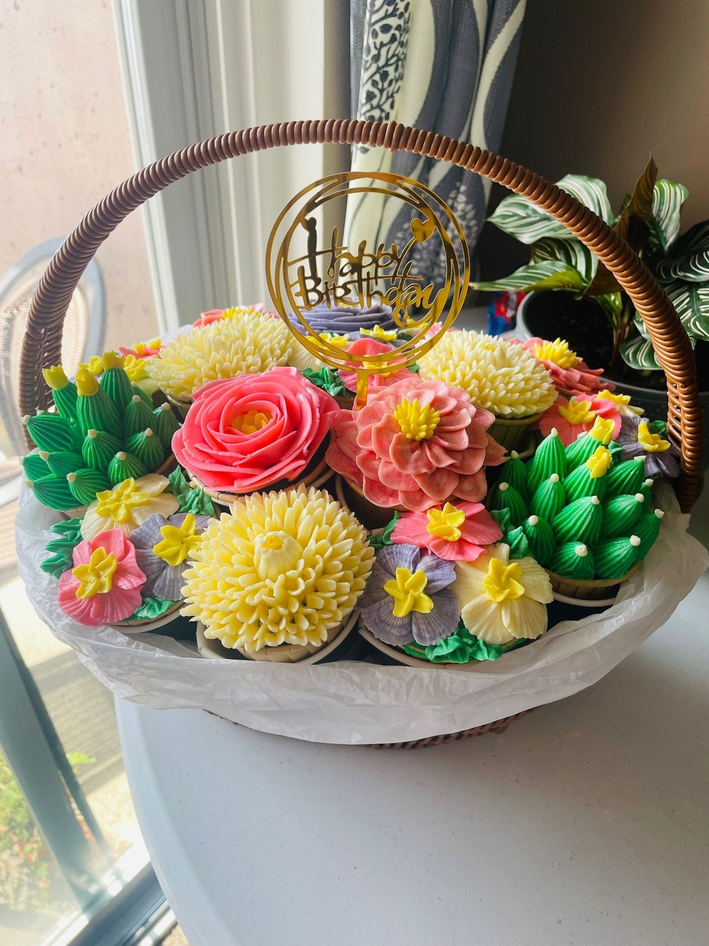 Basket of colorful flower-themed cupcakes with a 'Happy Birthday' topper by a window.