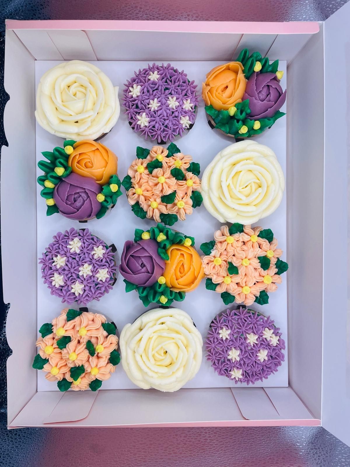 Box of colorful cupcakes decorated with intricately designed frosting flowers in various colors and patterns.