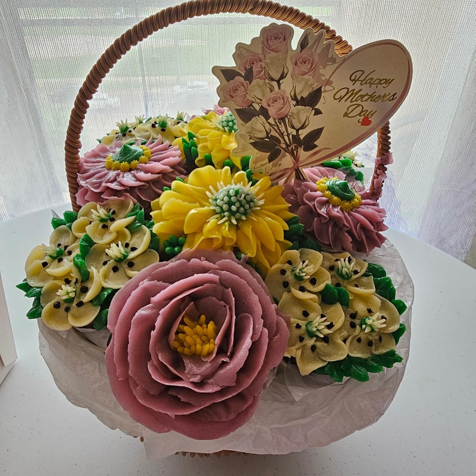 Basket of decorated cupcakes with flower designs and a card saying 'Happy Mother's Day' attached.